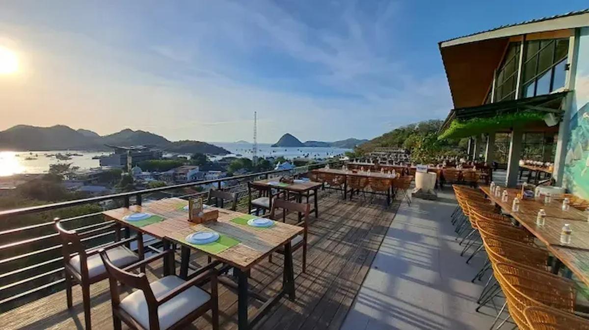 Restoran rooftop terbuka dengan meja kayu menghadap teluk dan pulau-pulau Labuan Bajo saat senja