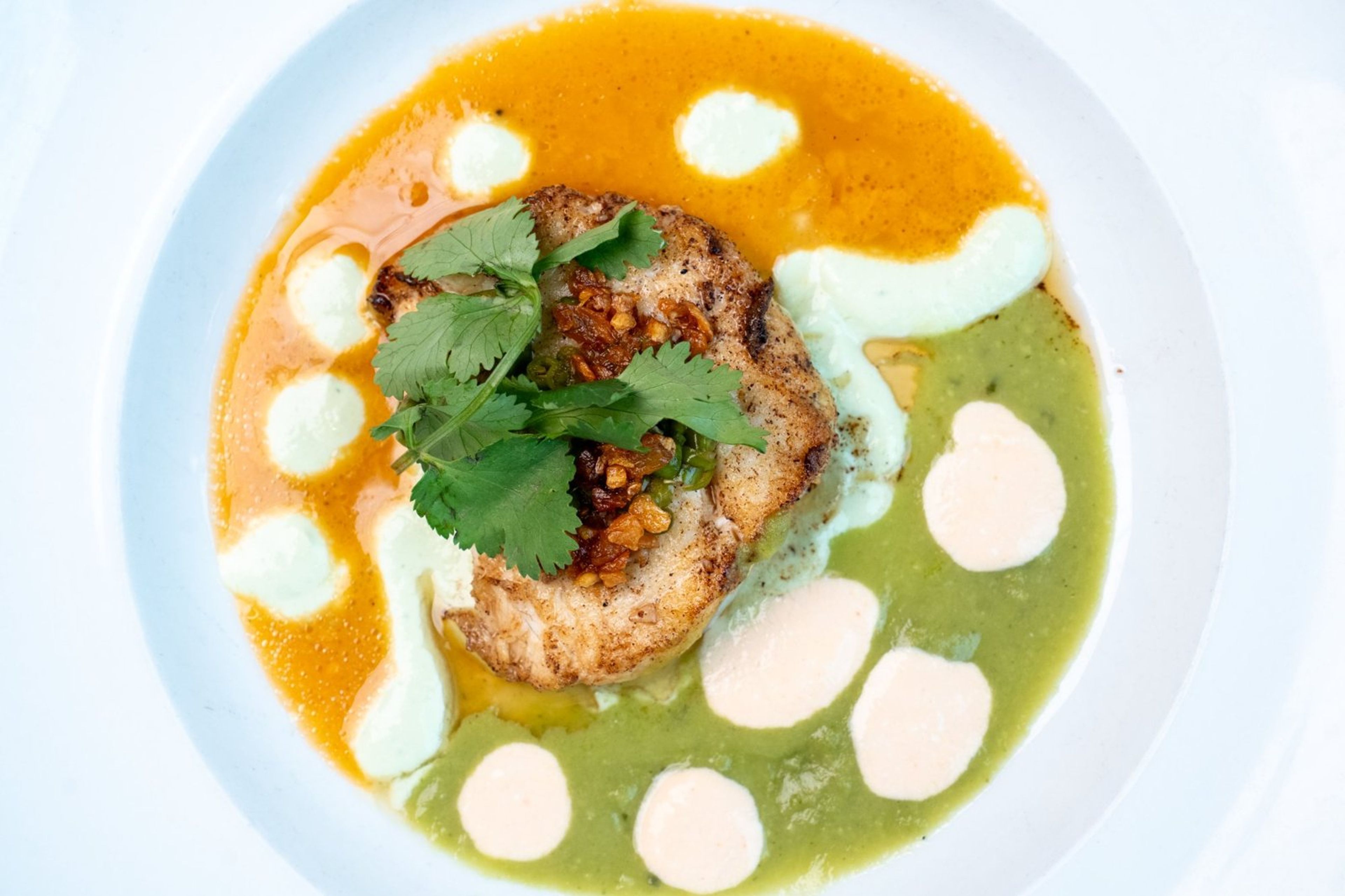 An overhead view of a plate with orange and green sauces, cilantro and dollops of cream