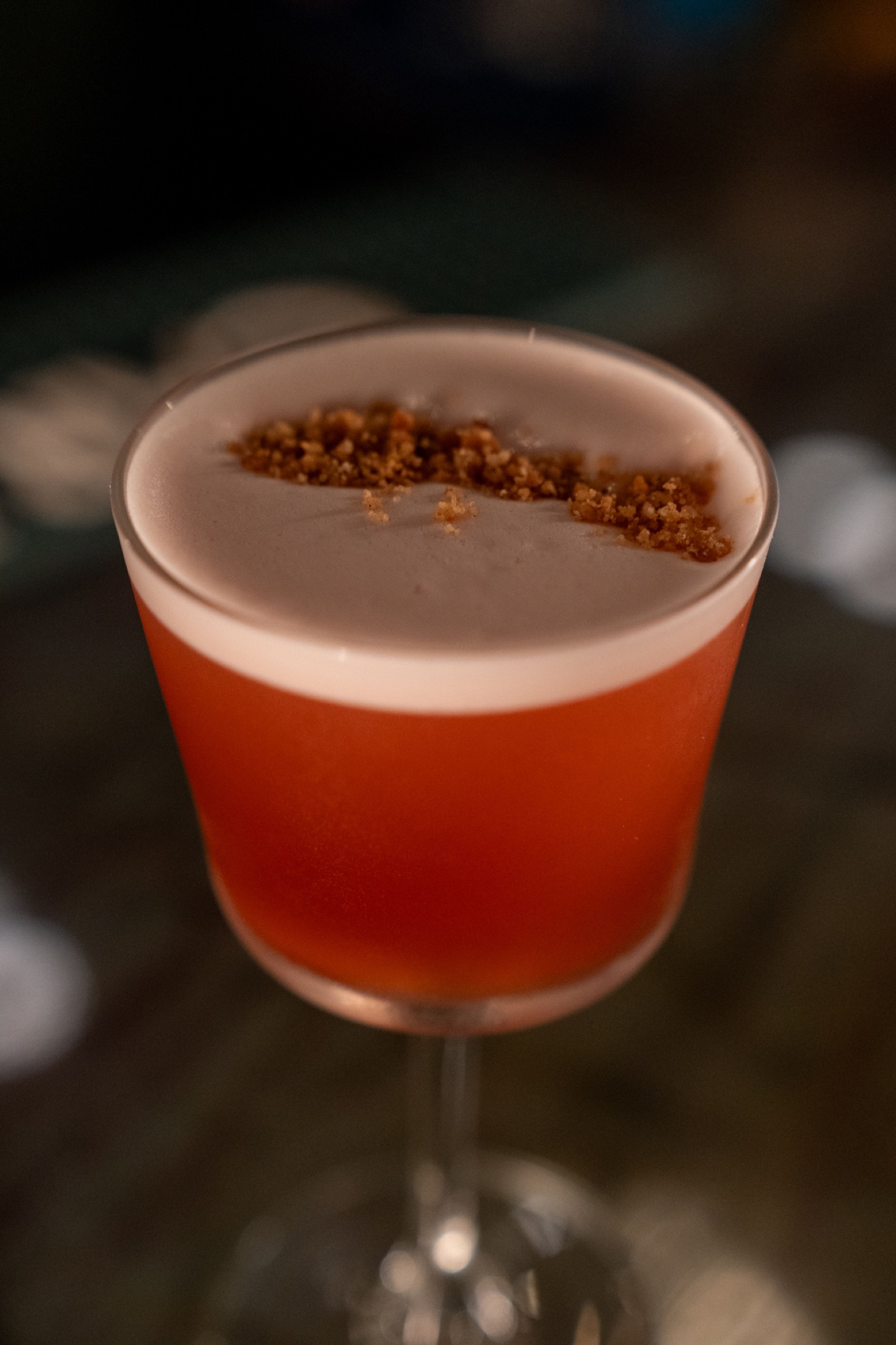 A red-orange cocktail topped with foam and a crumbled garnish