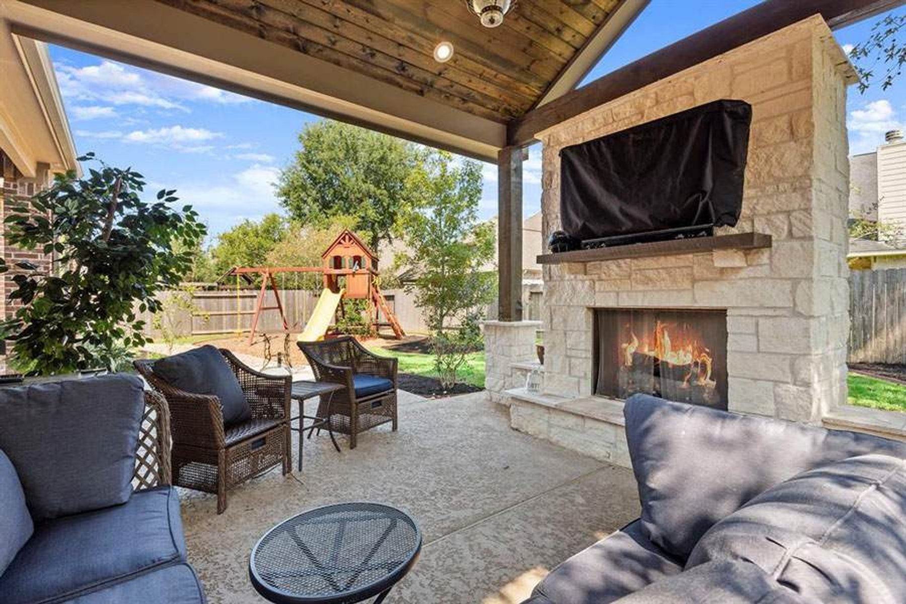 Covered patio with a built-in stone fireplace, outdoor seating, and a backyard playground