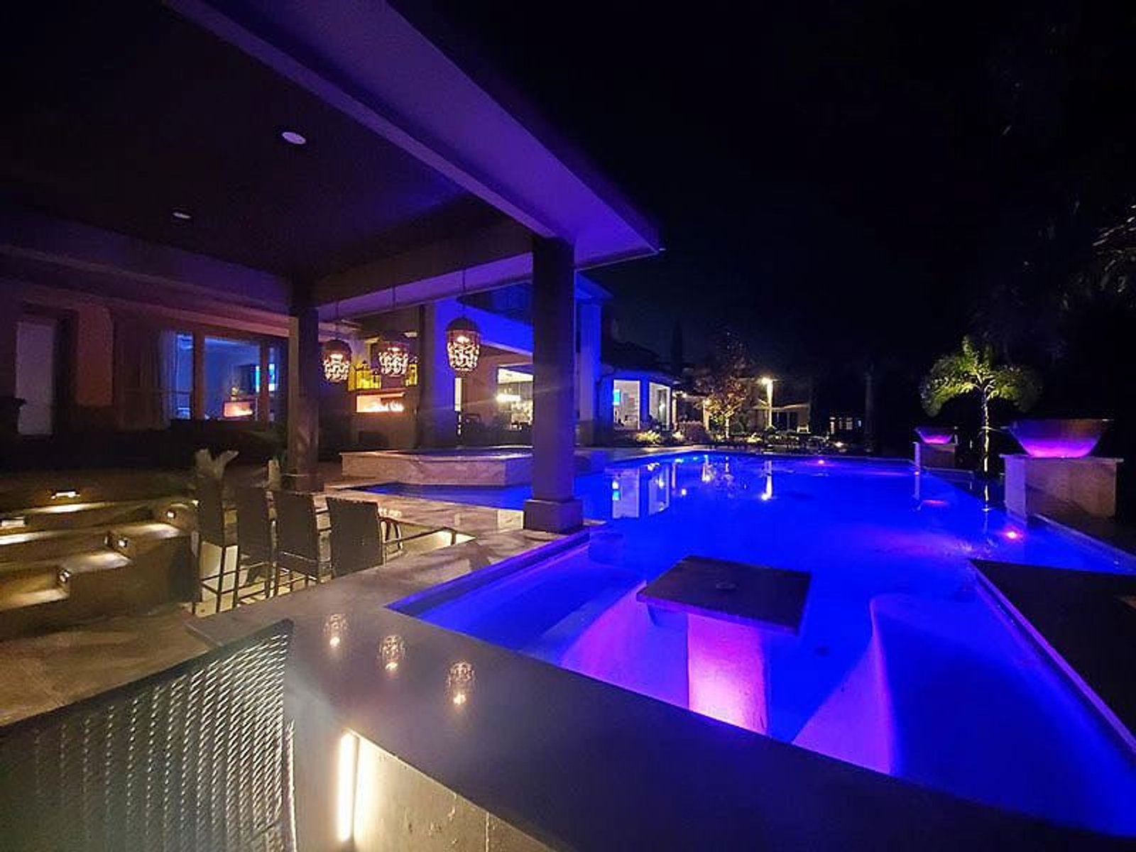 Luxury backyard swimming pool illuminated with vibrant purple lighting, featuring a spa and modern patio area