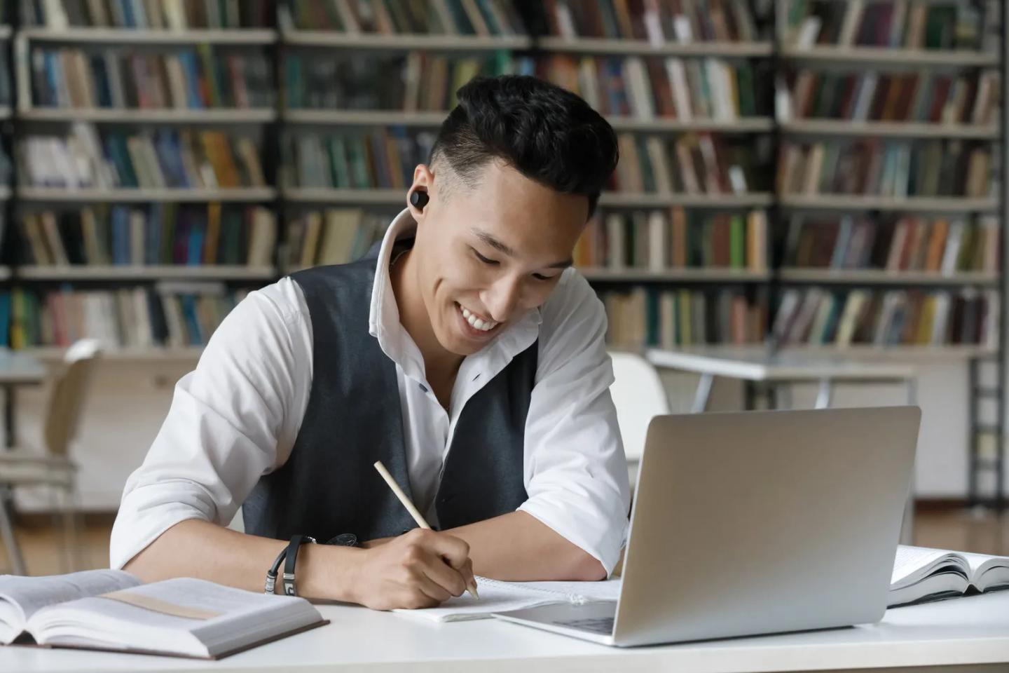 A student taking notes beside a laptop while studying in a library.