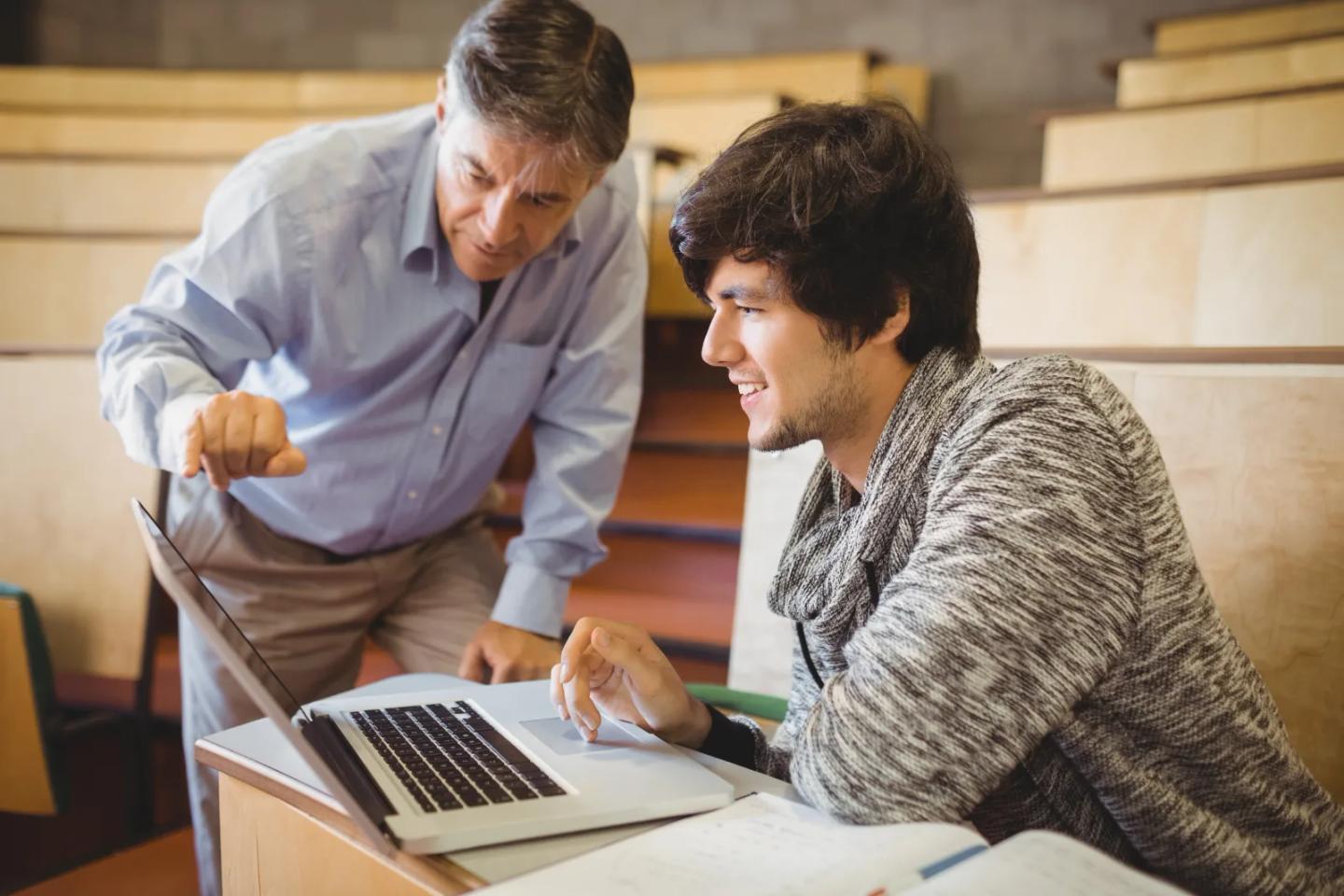 A professor helping a student at a laptop in a lecture hall.