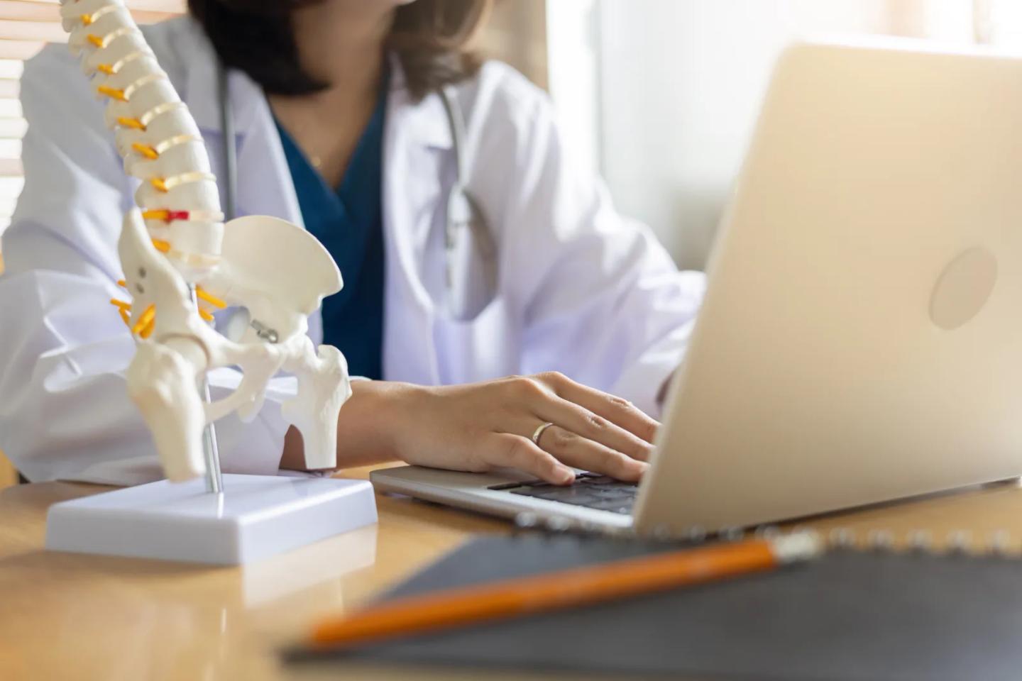 A physical therapy educator at a laptop beside an anatomical spine model.