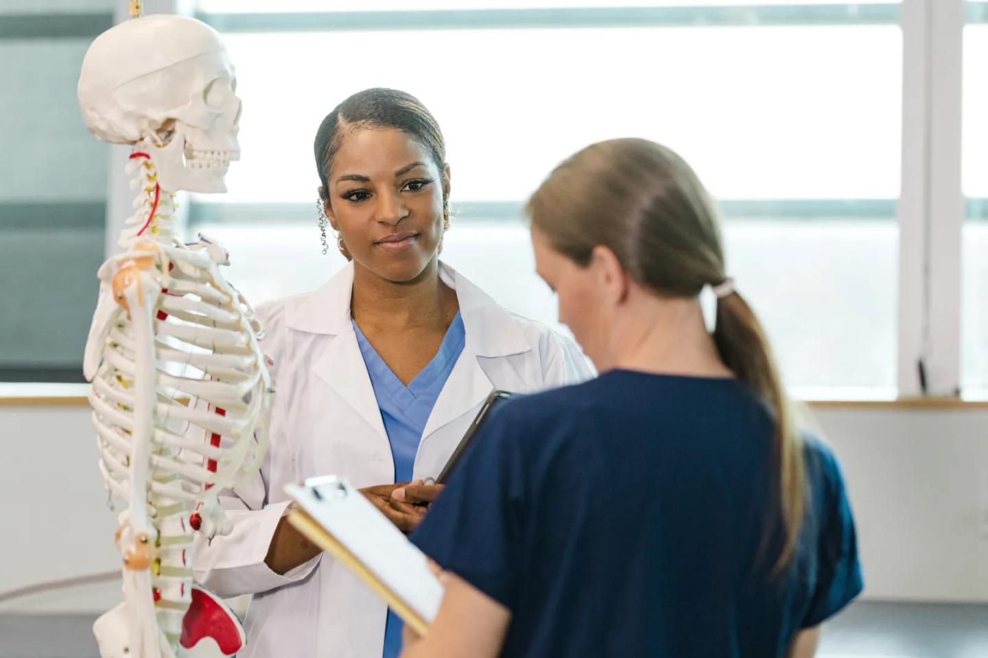 An instructor reviewing progress with a student beside an anatomy skeleton.