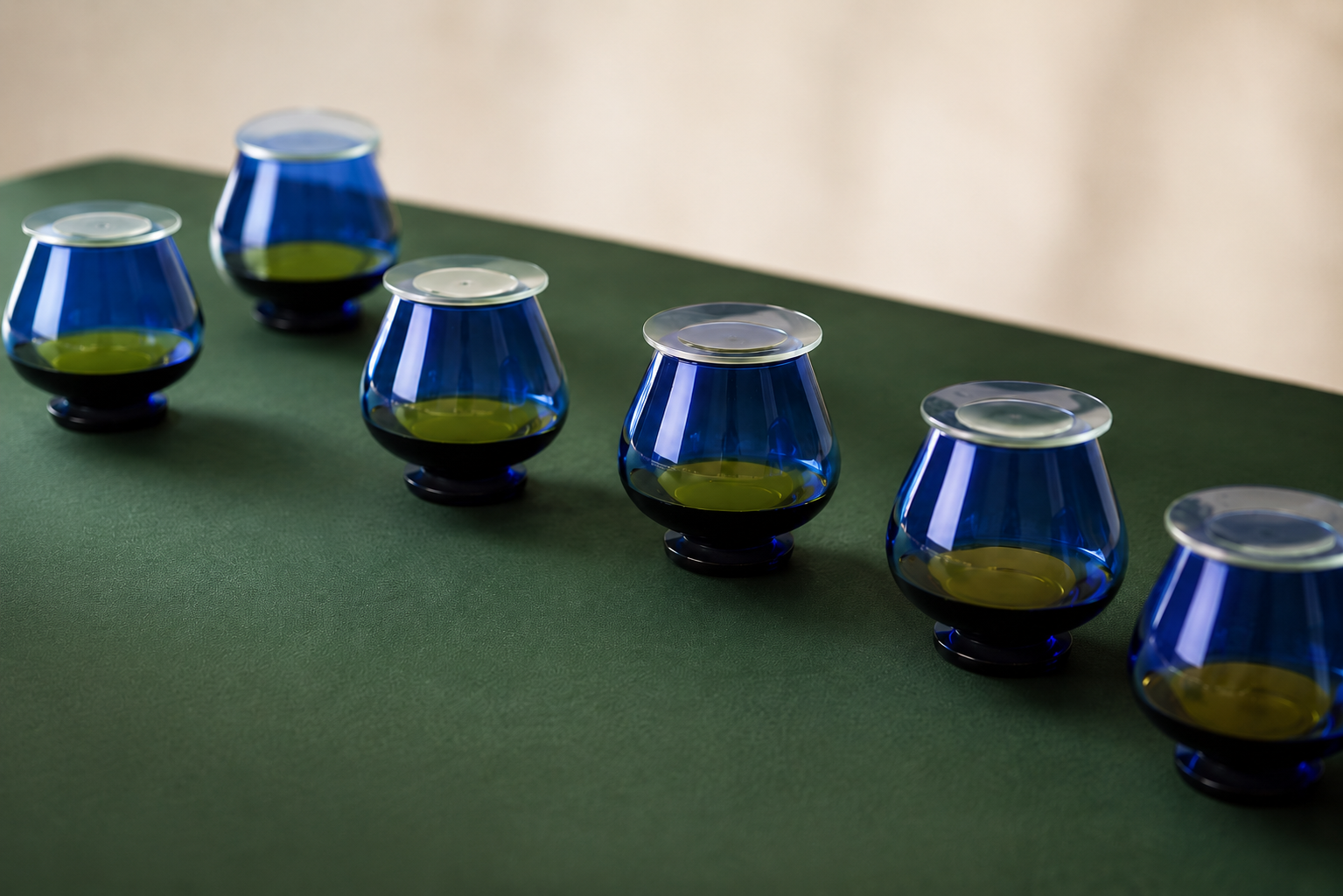 Six verres de dégustation d'huile d'olive bleu cobalt avec couvercles, remplis d'huile verte, sur une table vert foncé