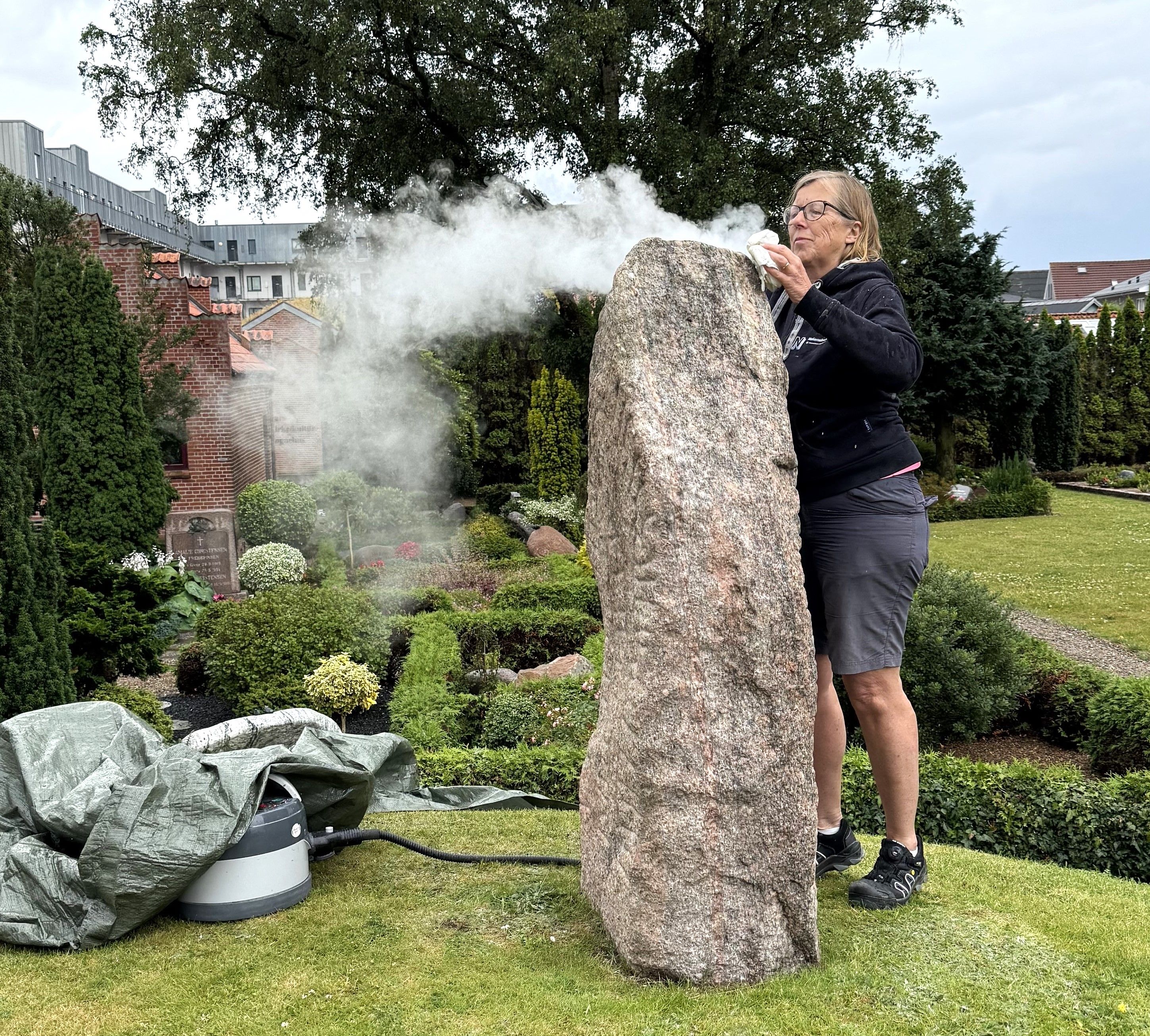 Kvindelig konservator afrenser runestenen udenfor ved Aars Kirke med damp.