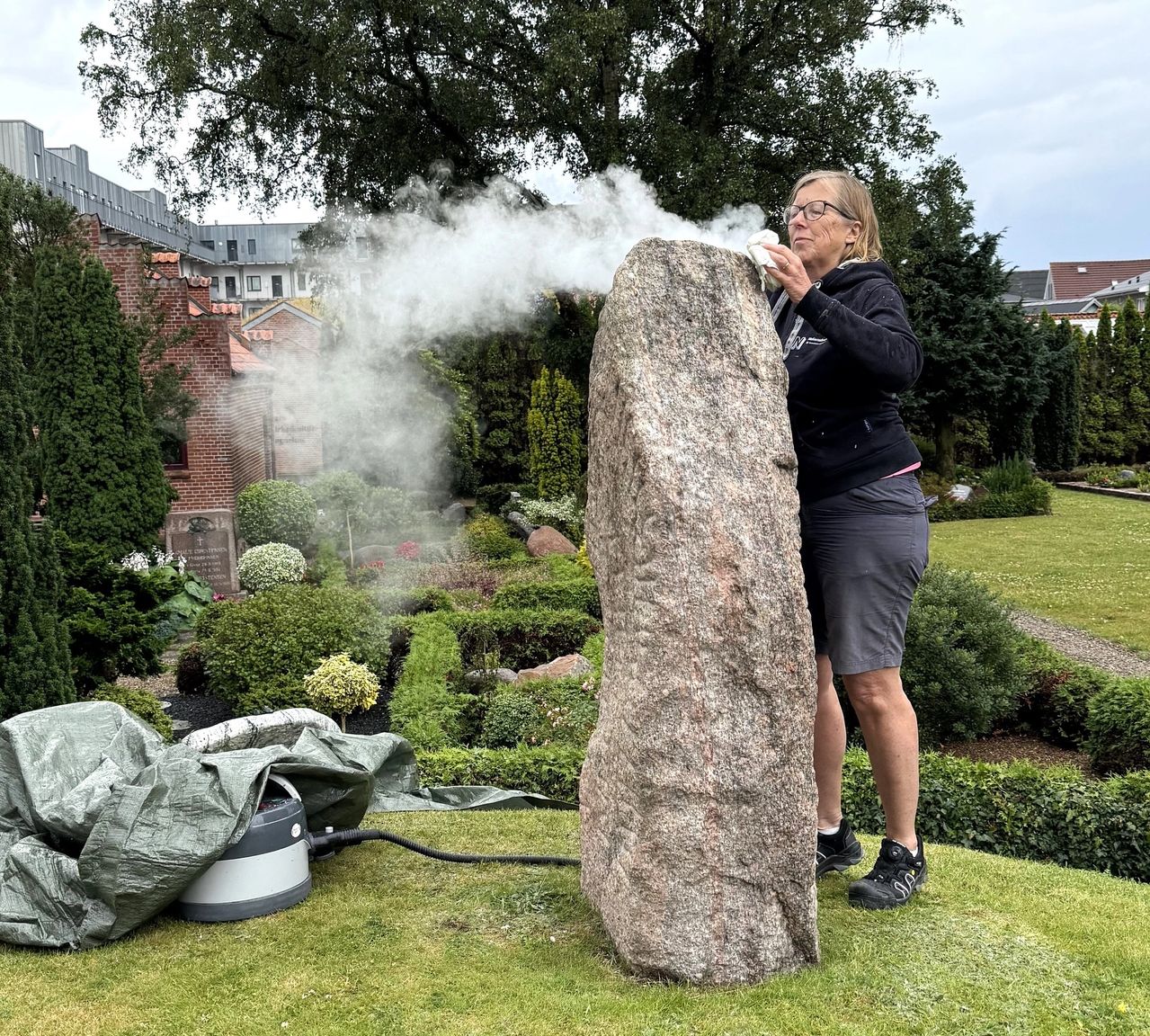 Kvindelig konservator afrenser runestenen udenfor ved Aars Kirke med damp.
