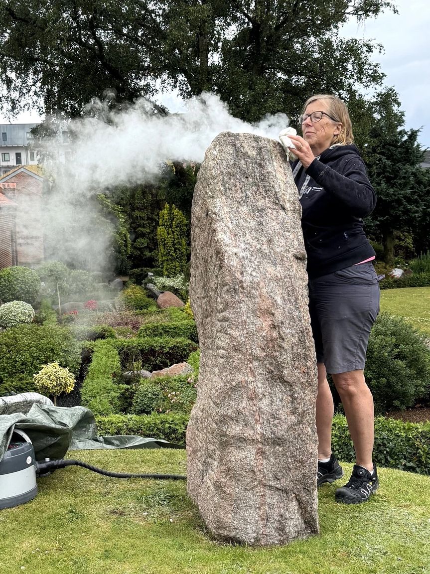 Kvindelig konservator afrenser runestenen udenfor ved Aars Kirke med damp.