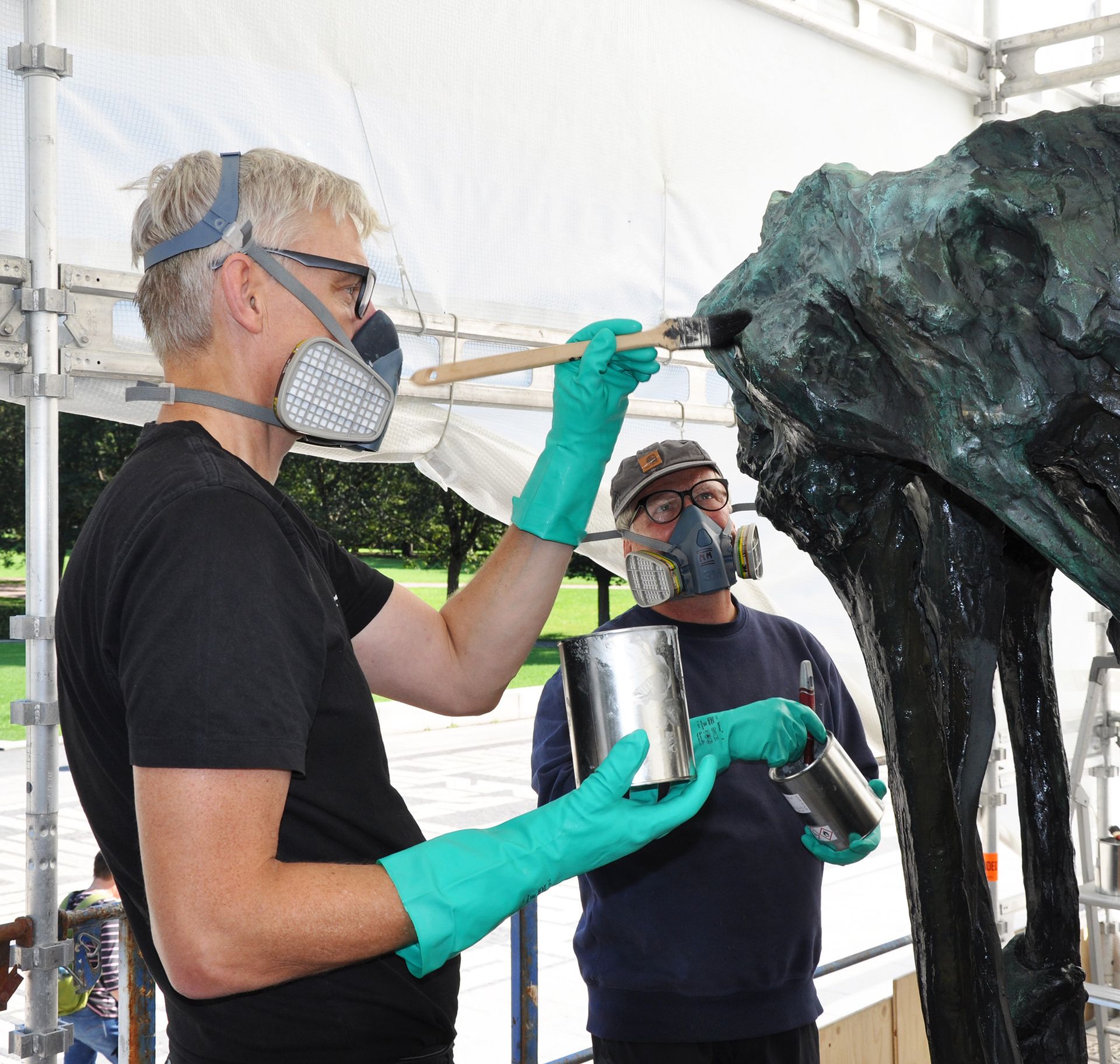 To mandlige konservatorer påfører voks på en metalskulptur, der forestiller et nøgent menneske, der hænger og holder om træets top. Skulpturen er fra Vigemuseet i Oslo, Norge, som står i deres park.