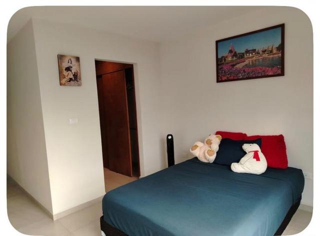 Recámara con cama vestida en azul, cojines rojos, paredes blancas y puerta de madera