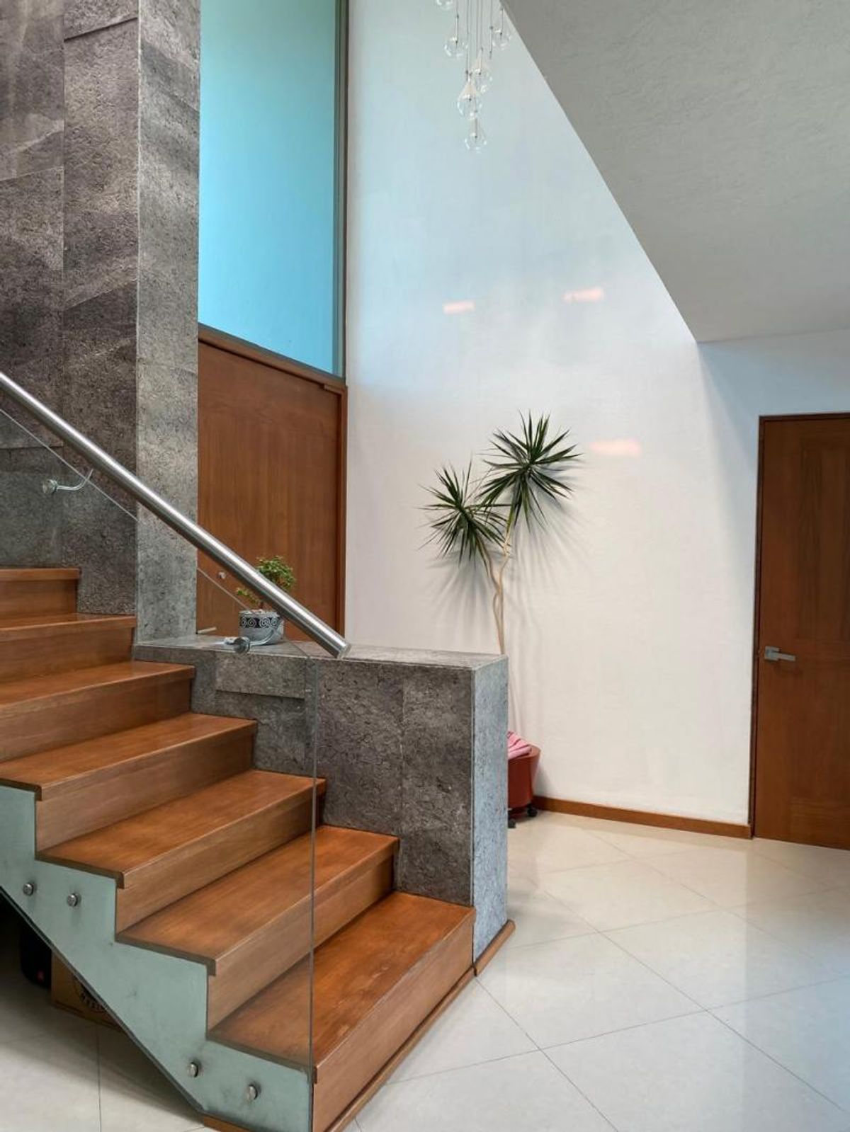 Escalera de madera con barandal de cristal, muro revestido en piedra y piso claro