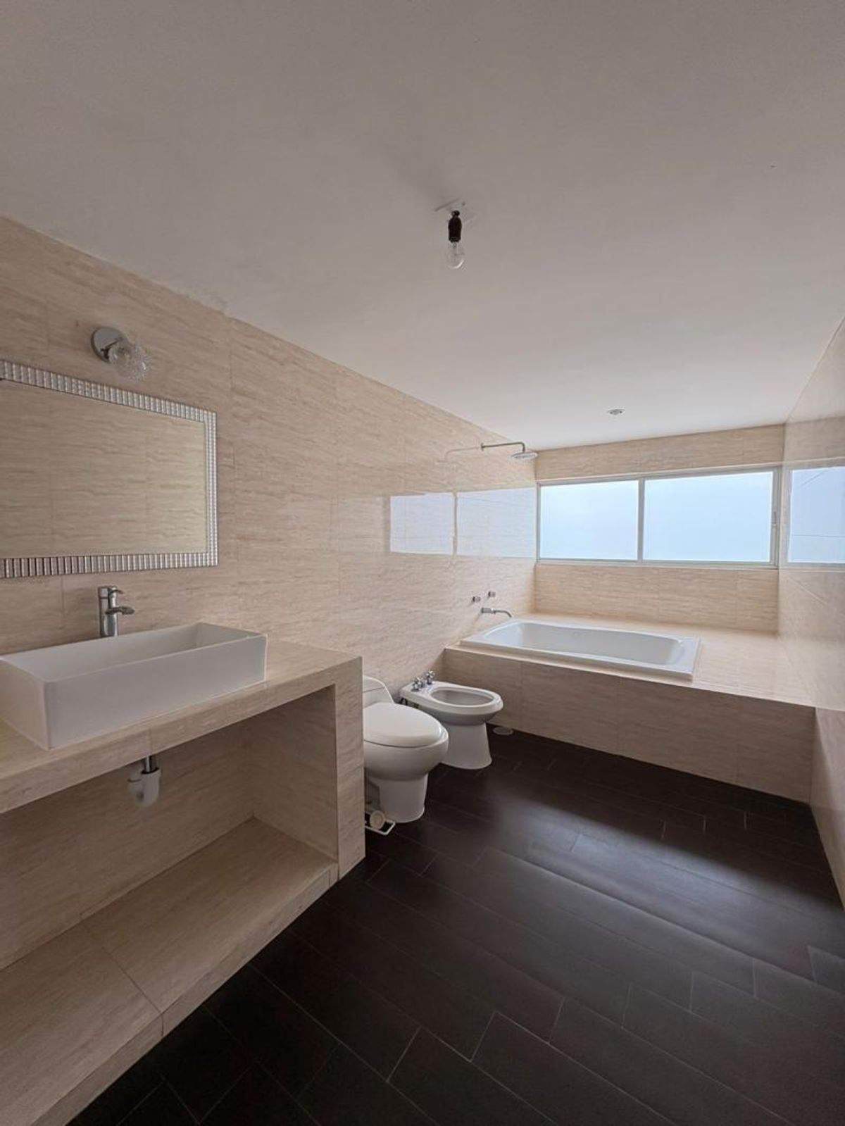 Baño con paredes de azulejo beige, lavabo blanco, inodoro, bidet, tina y piso oscuro