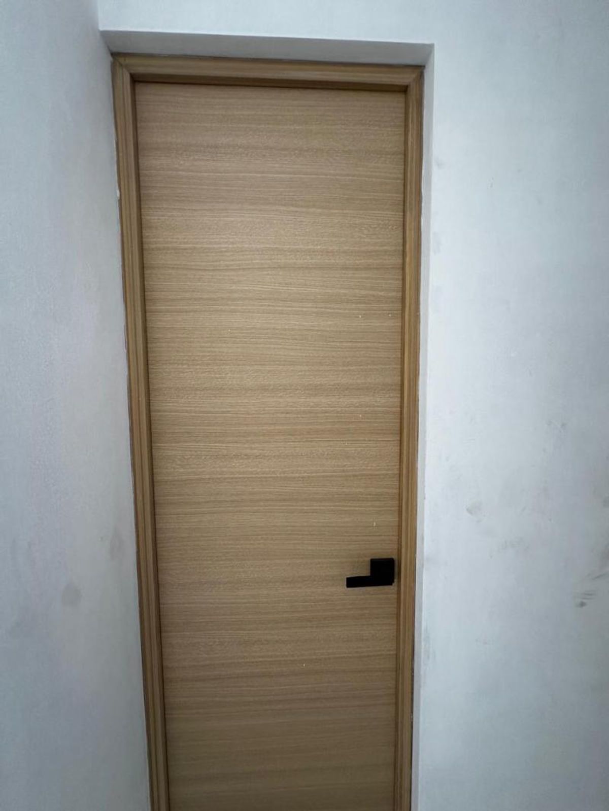 Puerta de madera clara con veteado horizontal, marco y manija negra sobre pared blanca
