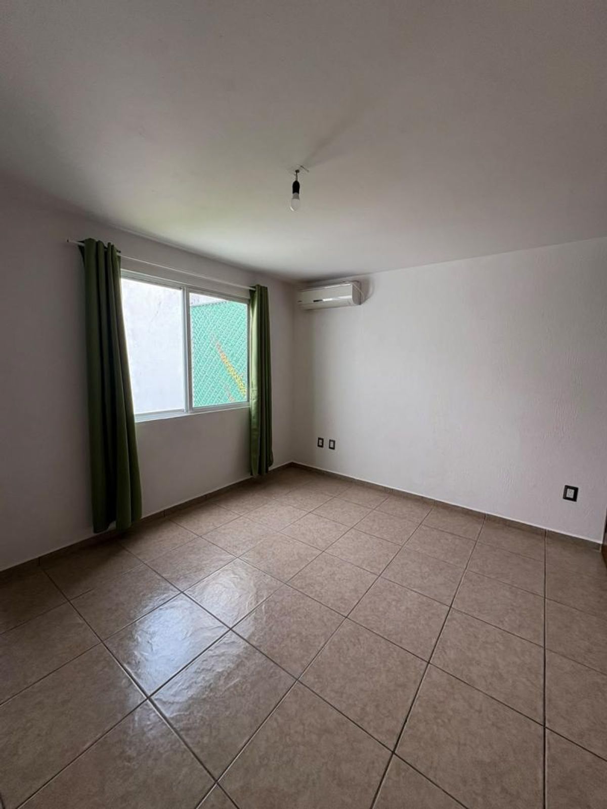 Habitación vacía con piso de cerámica beige, ventana con cortinas verdes y minisplit