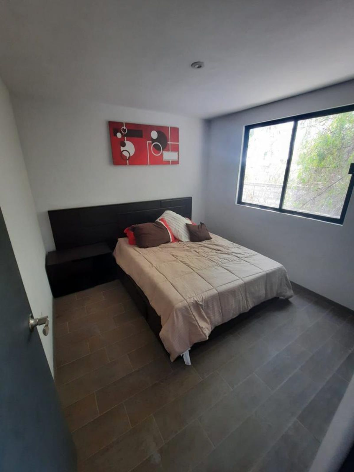Recámara con cama matrimonial, cabecera de madera oscura, ventana amplia y piso cerámico gris