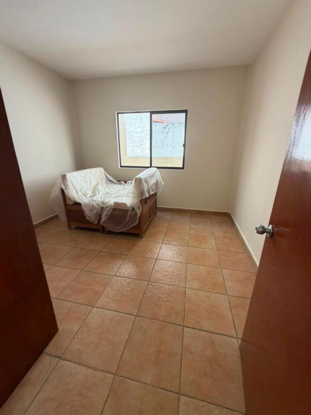Habitación con piso de cerámica terracota, ventana al fondo y sillón cubierto con plástico
