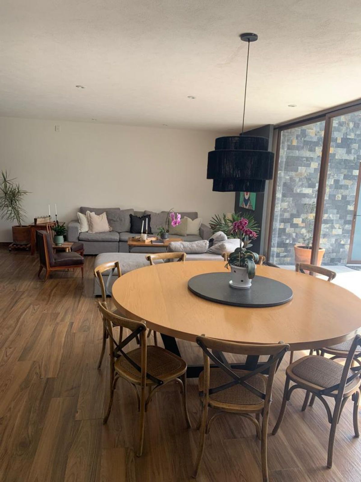 Comedor con mesa redonda de madera, lámpara colgante negra y sala al fondo