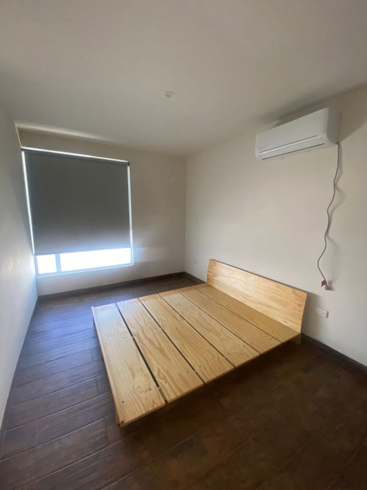 Recámara con base de cama de madera clara, piso tipo madera, persiana y minisplit