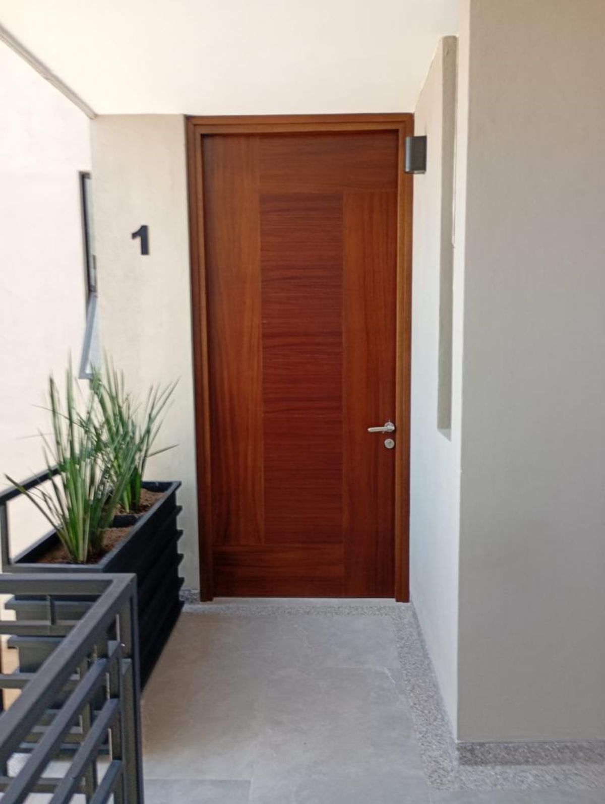 Acceso a departamento con puerta de madera, pared clara con número y jardinera con plantas