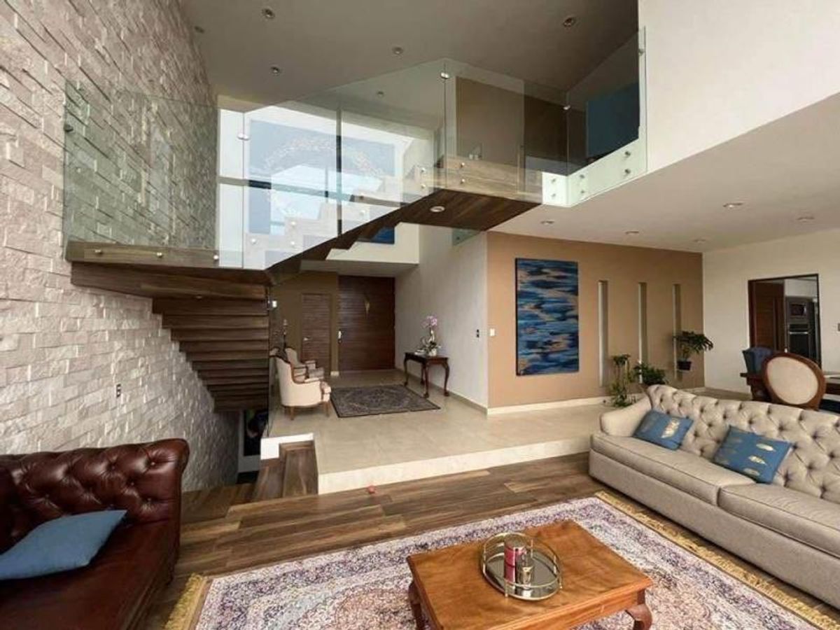Sala con sofás capitoné, tapete, muro de piedra y escalera con barandal de cristal