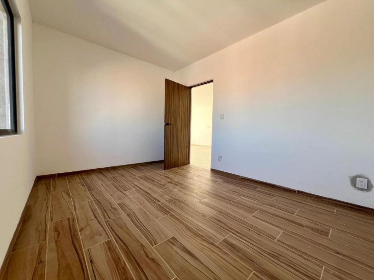 Habitación vacía con paredes blancas, piso cerámico tipo madera y puerta de madera abierta