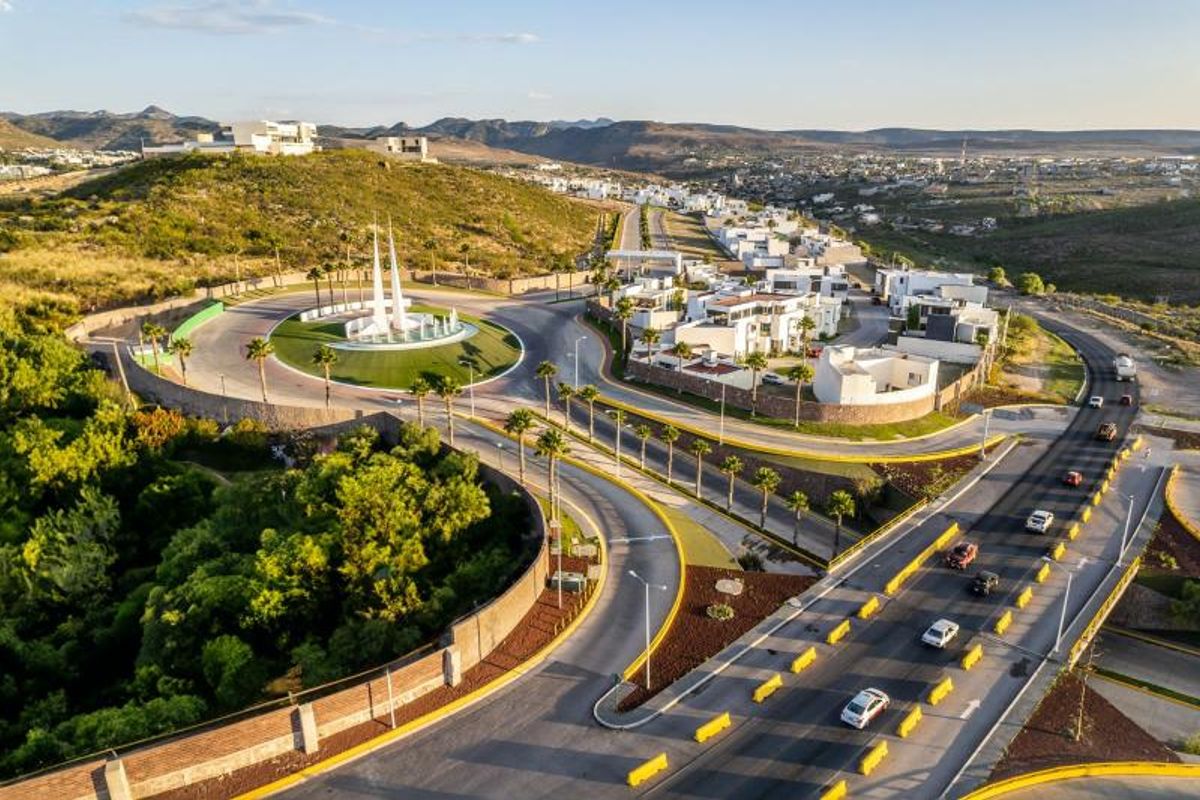 Fraccionamiento residencial con avenida, rotonda decorativa, palmeras, casas modernas y colinas de fondo