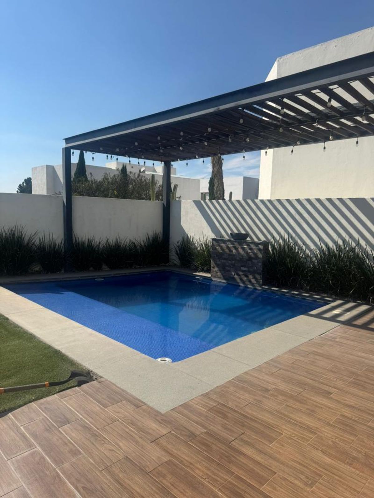 Alberca azul rodeada de piso cerámico tipo madera y pérgola metálica con luces colgantes
