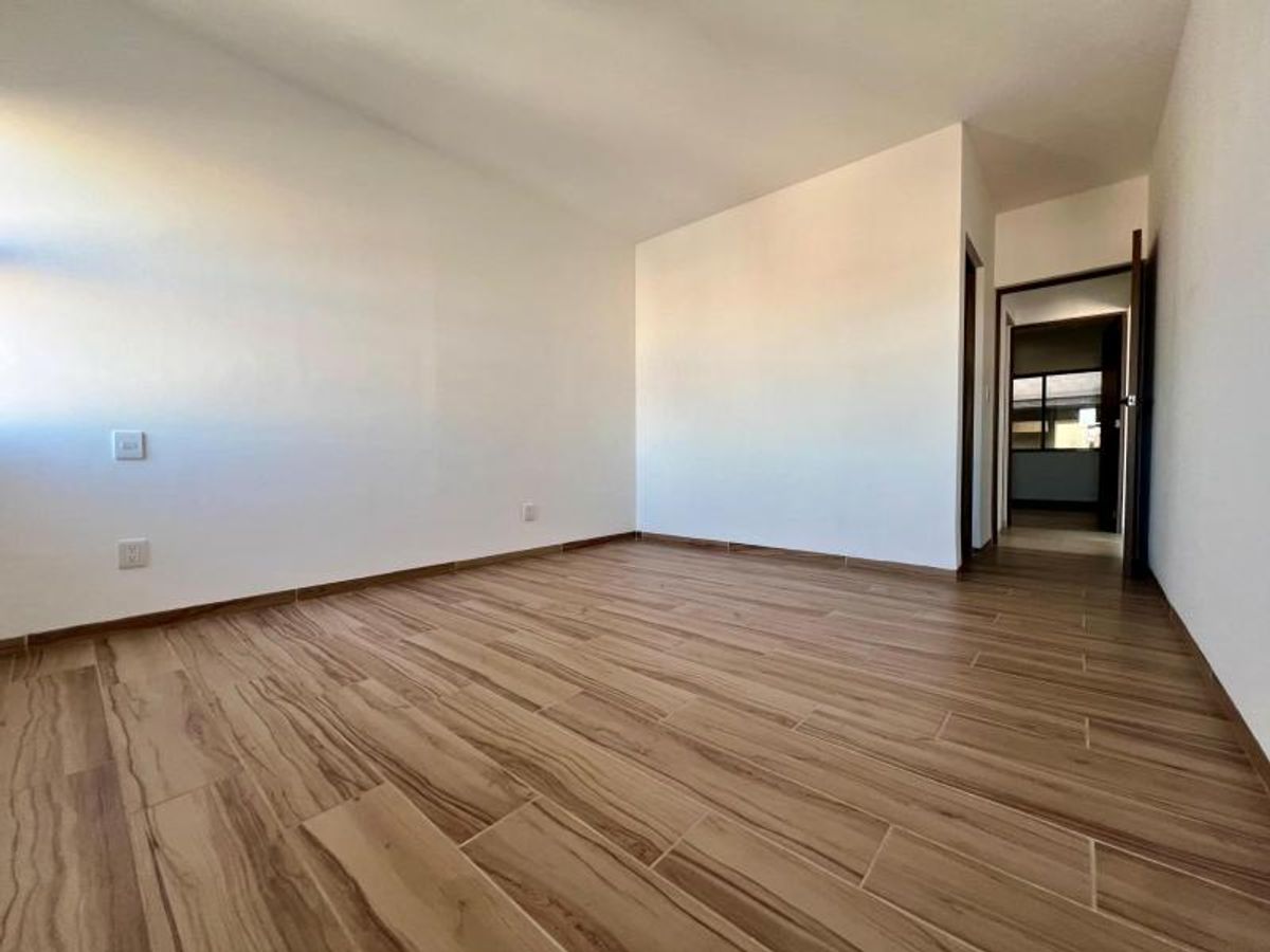 Habitación vacía con paredes blancas, piso cerámico aspecto madera y acceso a un pasillo