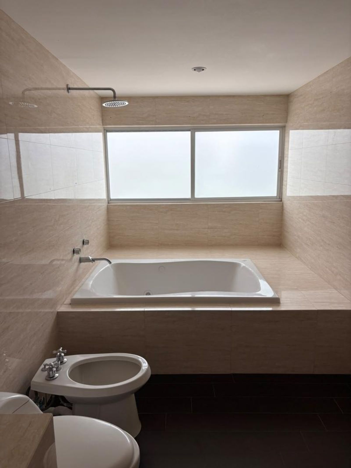 Baño con tina empotrada, regadera superior, ventana esmerilada y paredes revestidas de azulejos beige