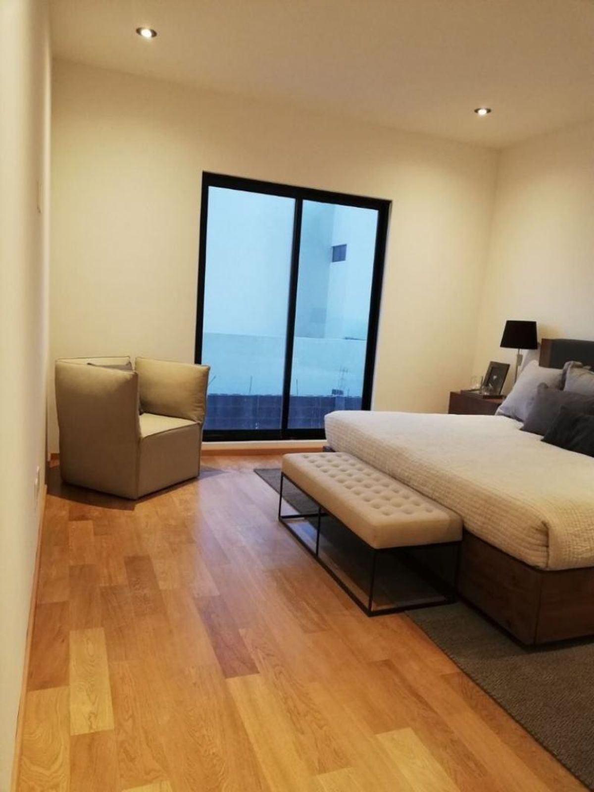 Recámara con piso de madera, cama con banca tapizada, sillón contemporáneo y gran ventanal