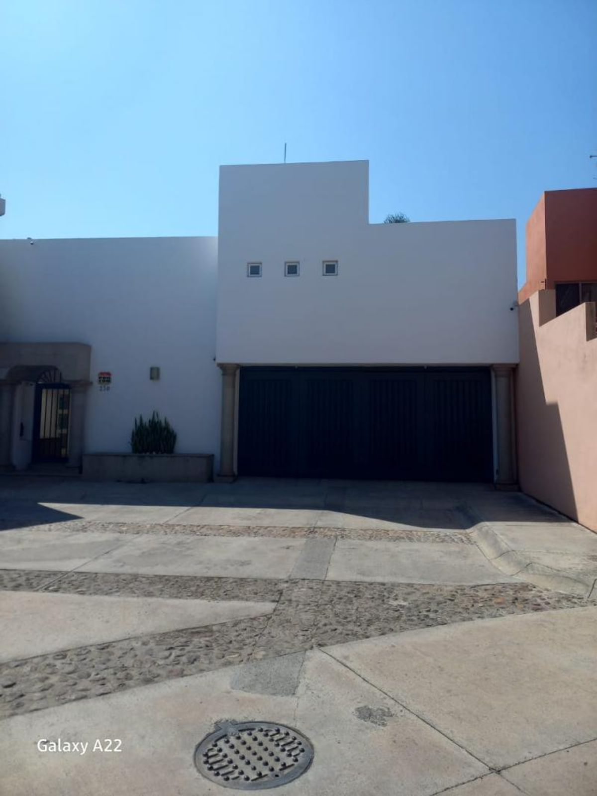 Fachada blanca con portón negro, tres ventanas pequeñas y piso de concreto empedrado