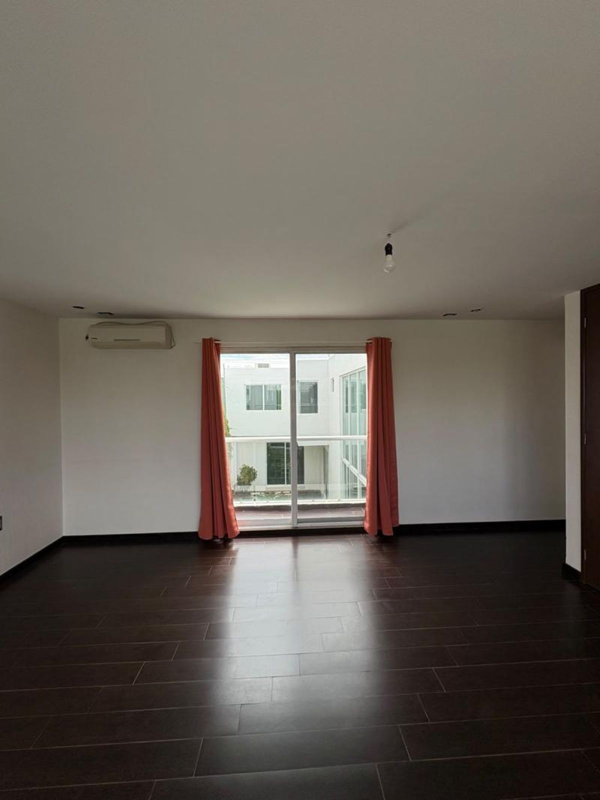 Habitación vacía con piso oscuro, paredes blancas, aire acondicionado y ventanal con cortinas salmón
