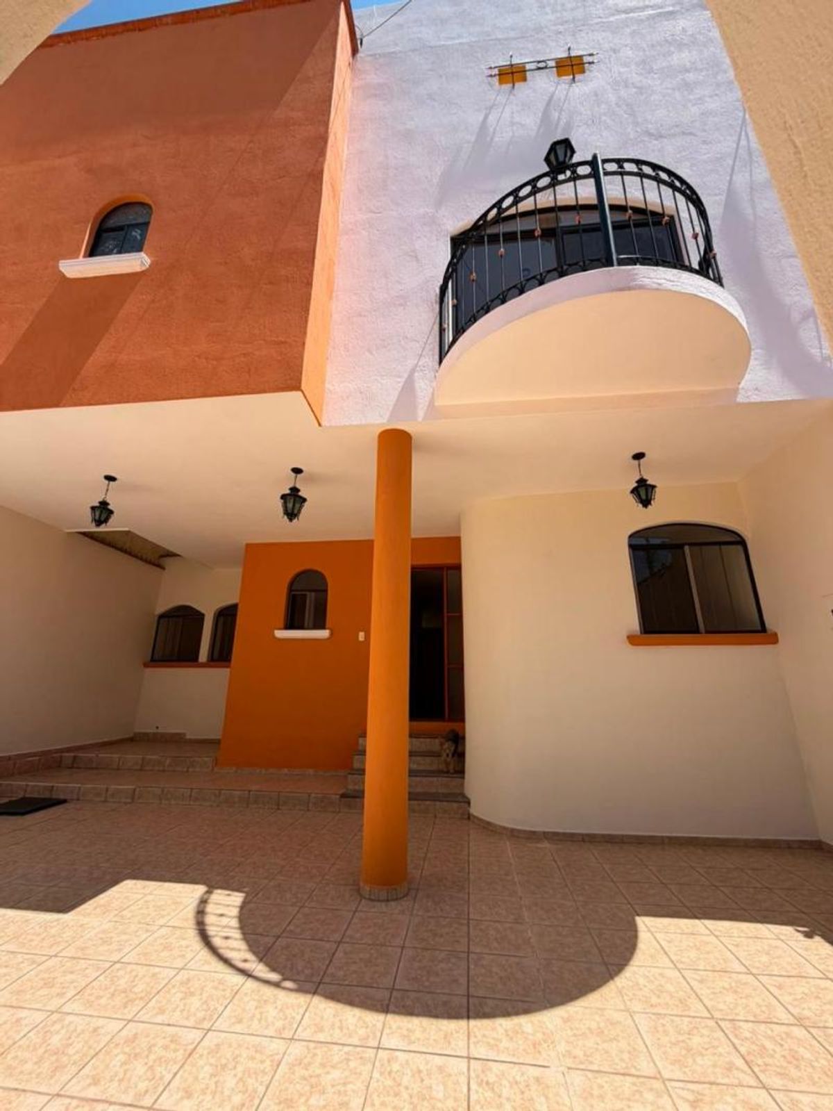 Patio con piso cerámico, columna central naranja, fachada bicolor y balcón con herrería negra