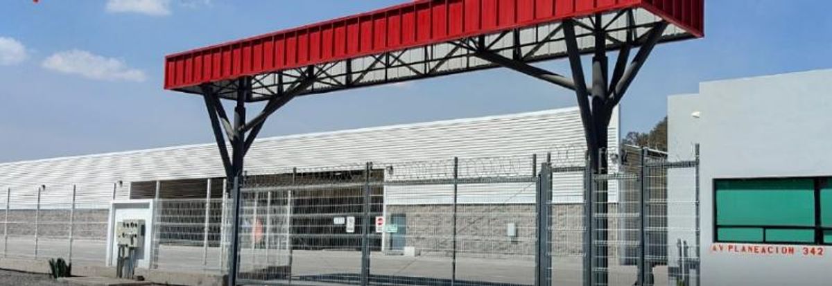 Entrada de parque industrial con estructura metálica roja, reja de seguridad y fachada blanca
