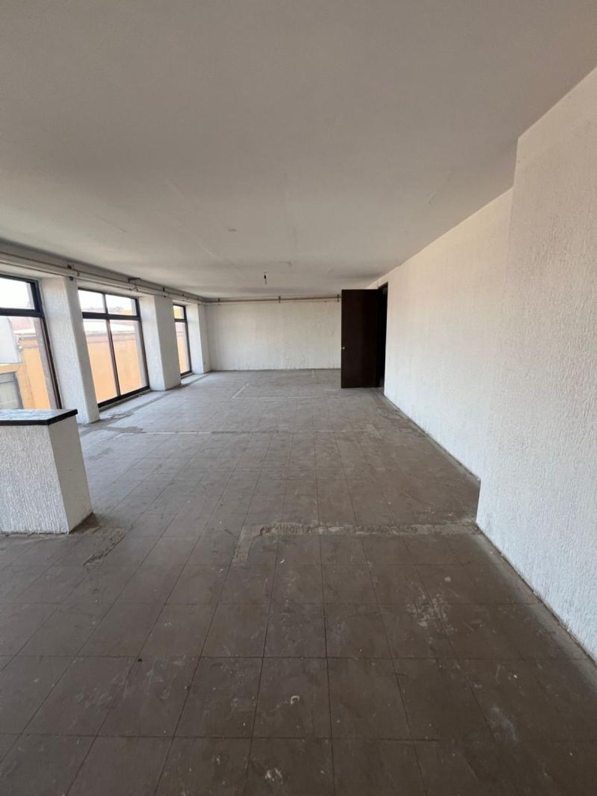 Espacio interior vacío con piso de baldosas grises, paredes blancas y ventanales laterales