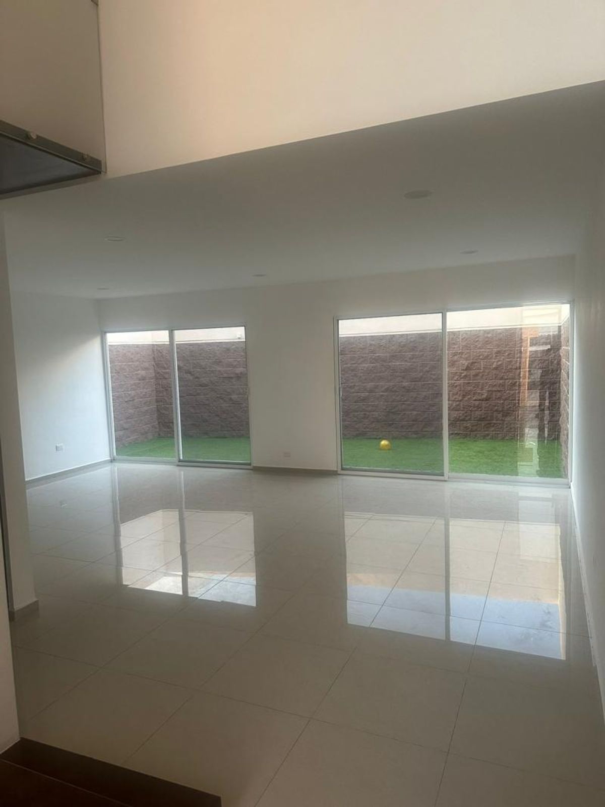 Sala vacía con piso de loseta brillante y ventanales hacia patio con césped sintético