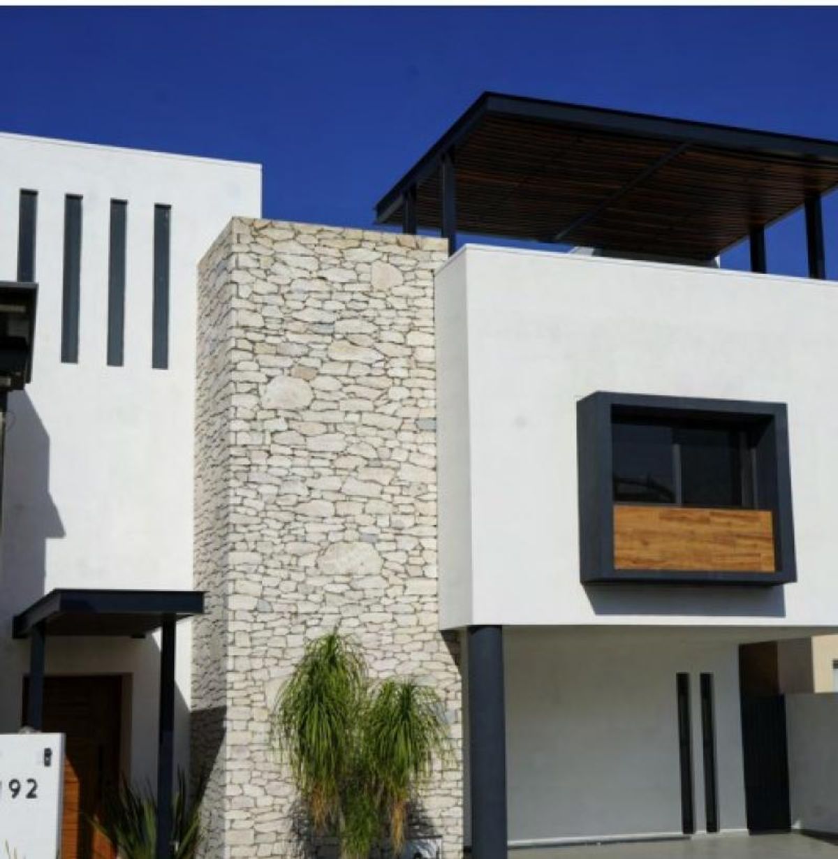 Fachada moderna blanca con muro de piedra, marco negro en ventana y pérgola superior