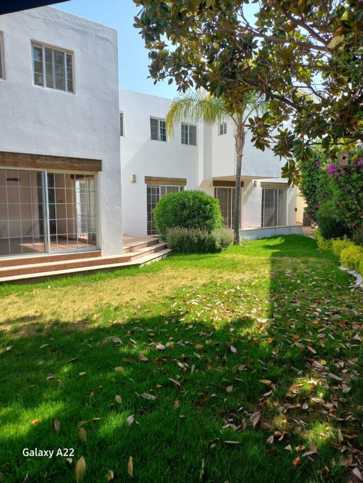 Fachada blanca de dos pisos con ventanales corredizos y jardín verde con árboles