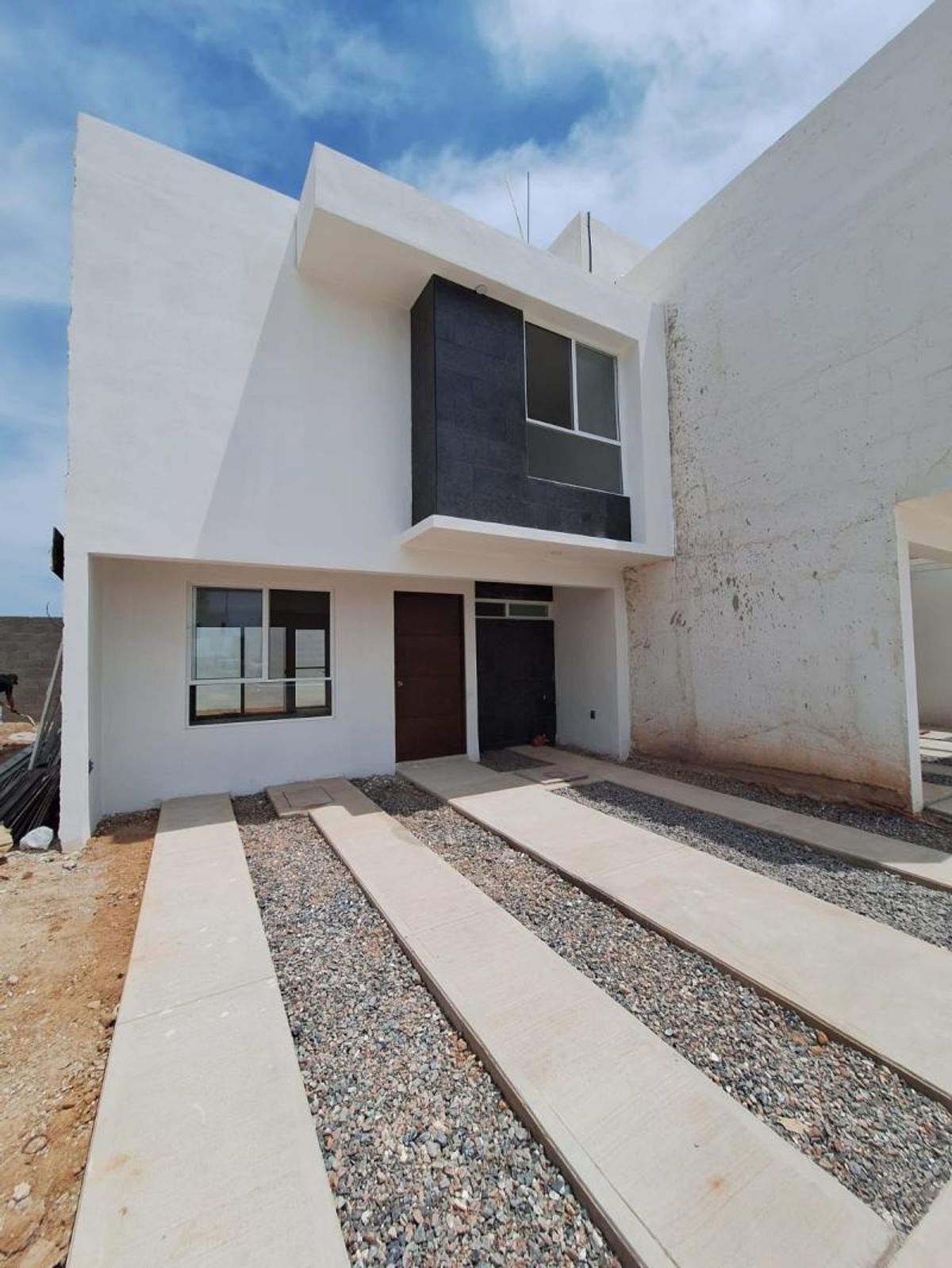 Fachada blanca de dos niveles con puerta oscura y huellas de concreto sobre grava