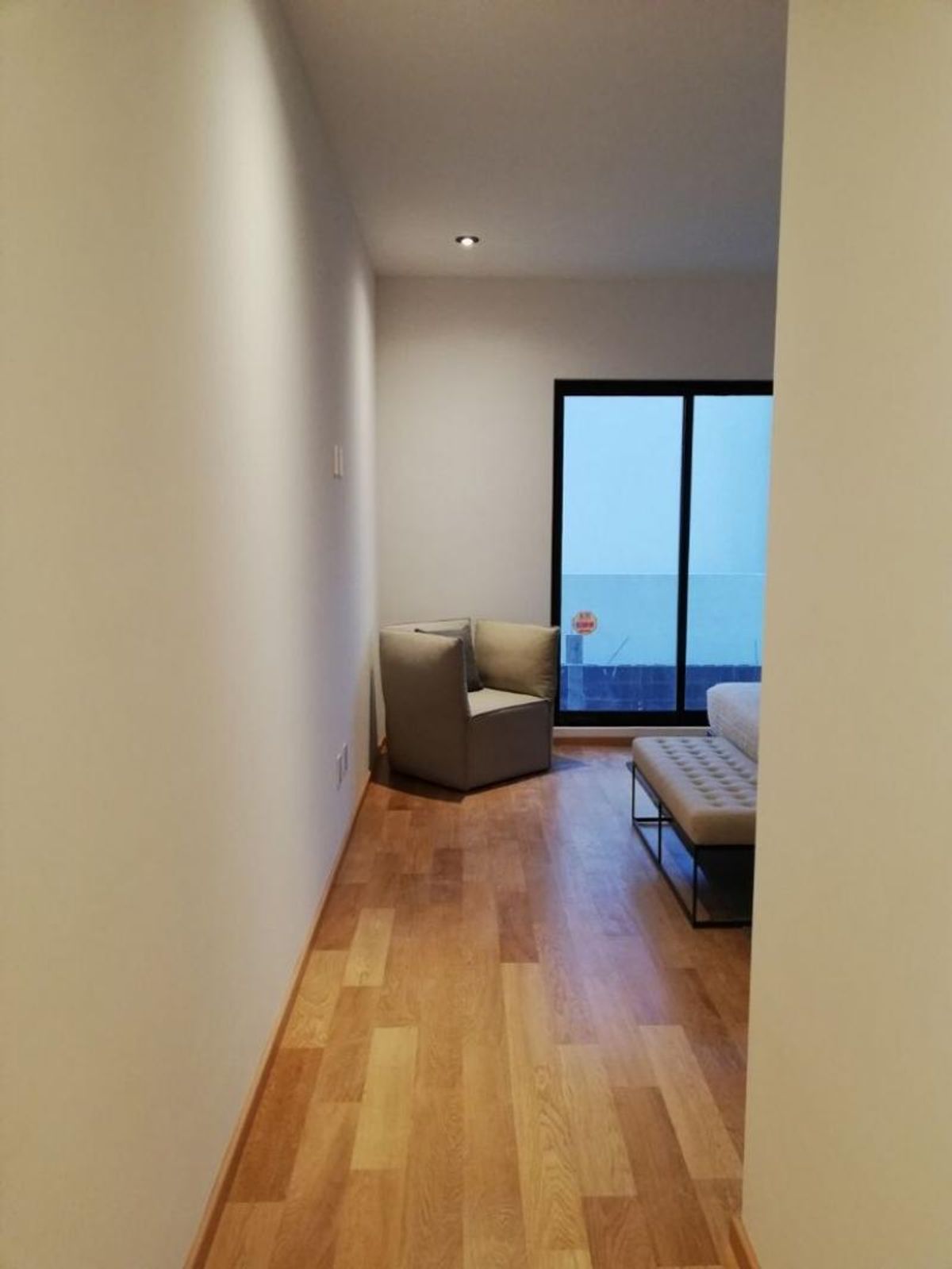 Pasillo con piso de madera, sillón individual gris al fondo, banca tapizada y ventanal