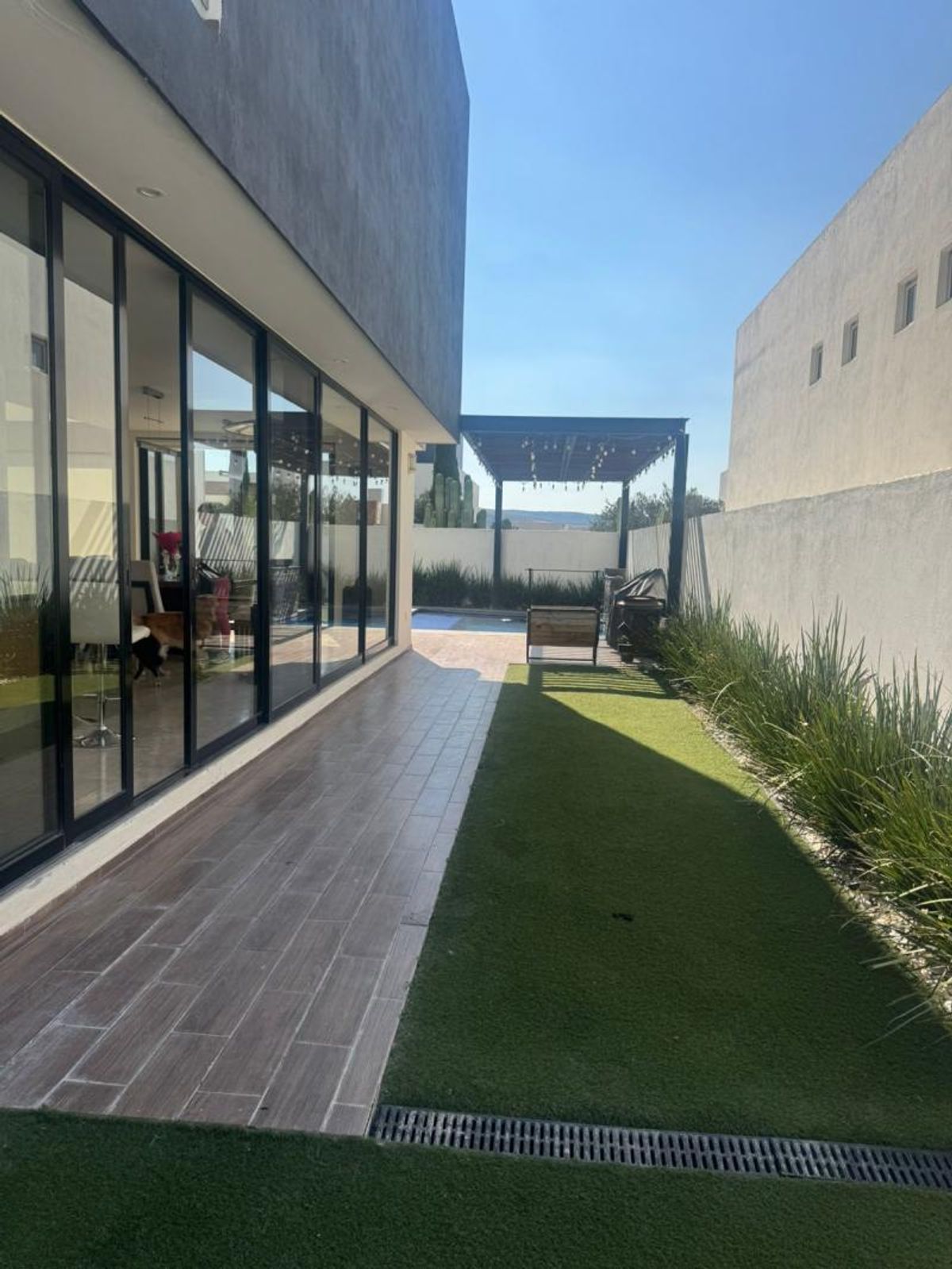Patio con piso de tipo madera, pasto sintético, grandes ventanales y pérgola al fondo