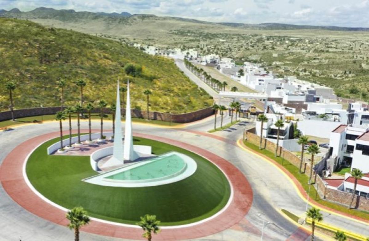 Glorieta de entrada con dos estructuras blancas, fuente central, palmeras y casas residenciales
