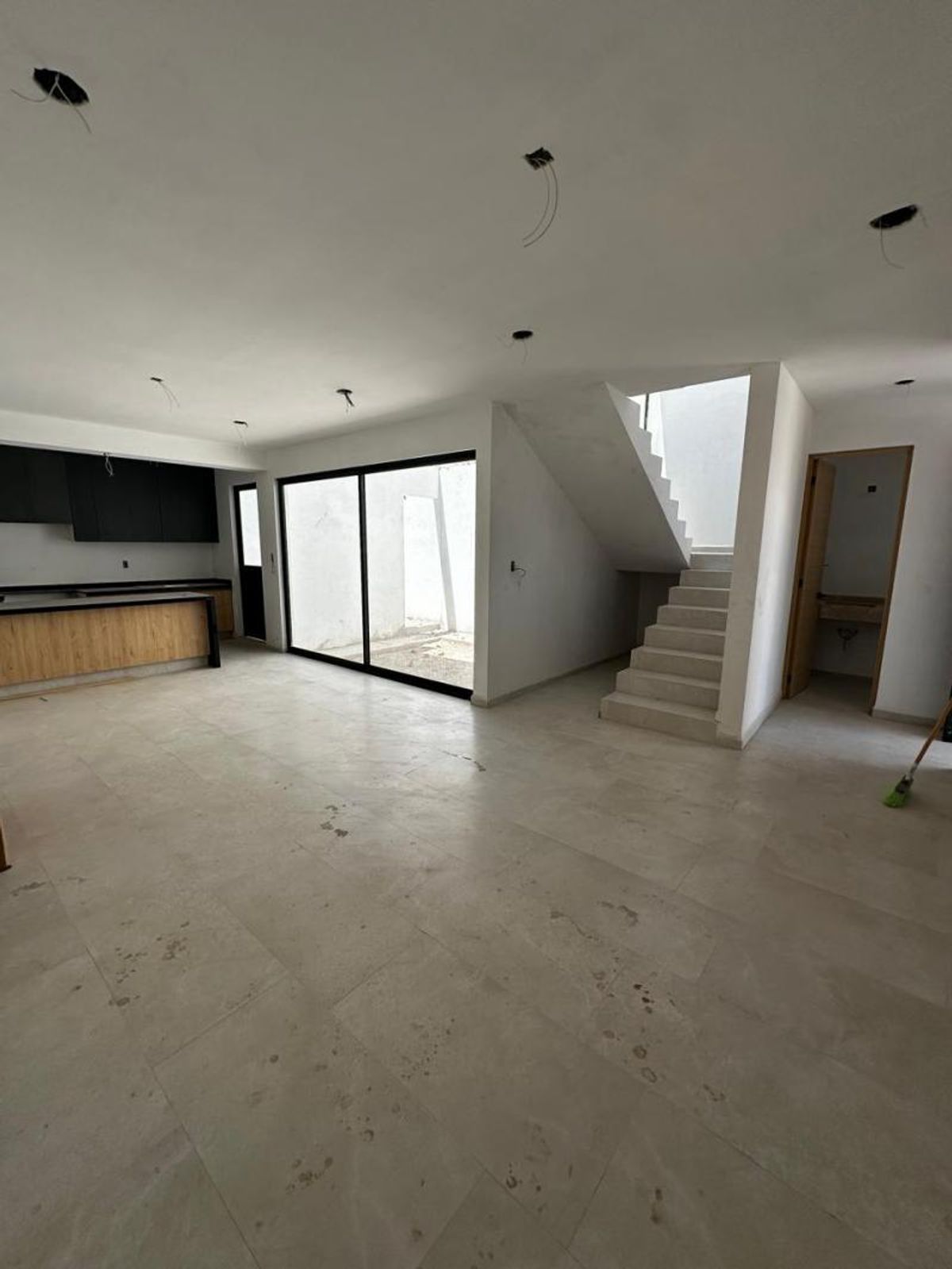 Espacio abierto con piso claro, cocina al fondo, ventanal de marco negro y escalera