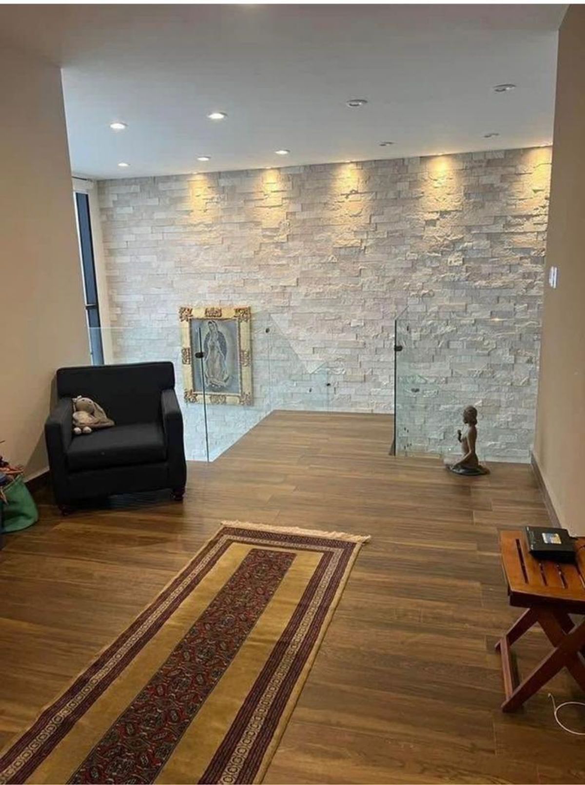 Vestíbulo con piso de madera, muro de piedra decorativa iluminado y barandal de vidrio