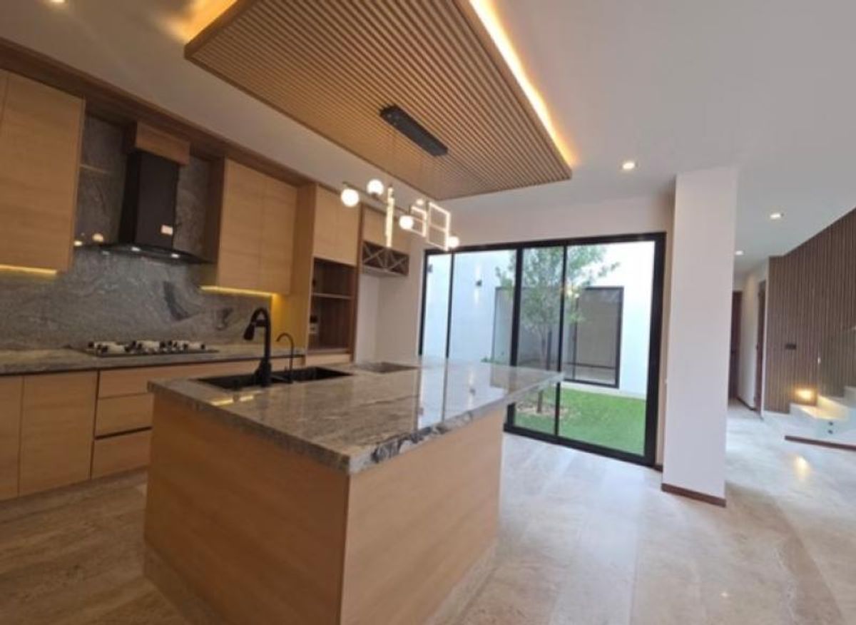 Cocina con isla de granito, gabinetes de madera, plafón iluminado y ventanales al jardín