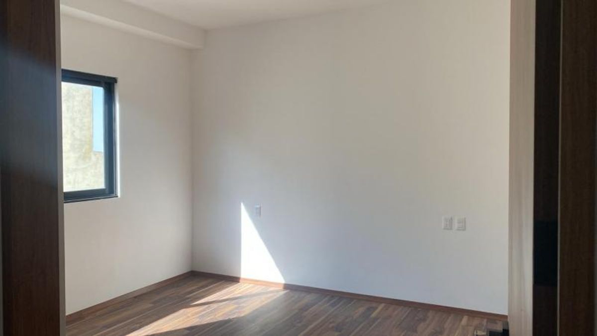 Habitación vacía con piso de madera oscura, paredes blancas y ventana con luz natural
