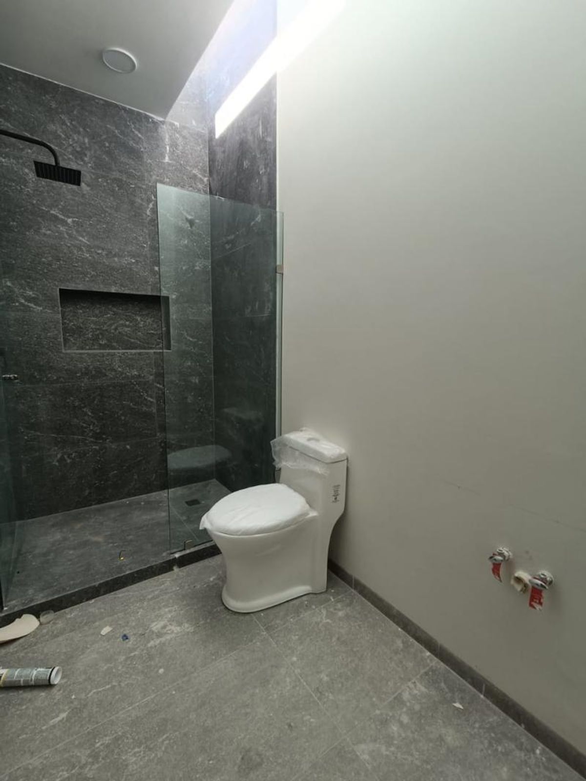 Baño con sanitario blanco, cancel de cristal y muro de regadera en revestimiento gris
