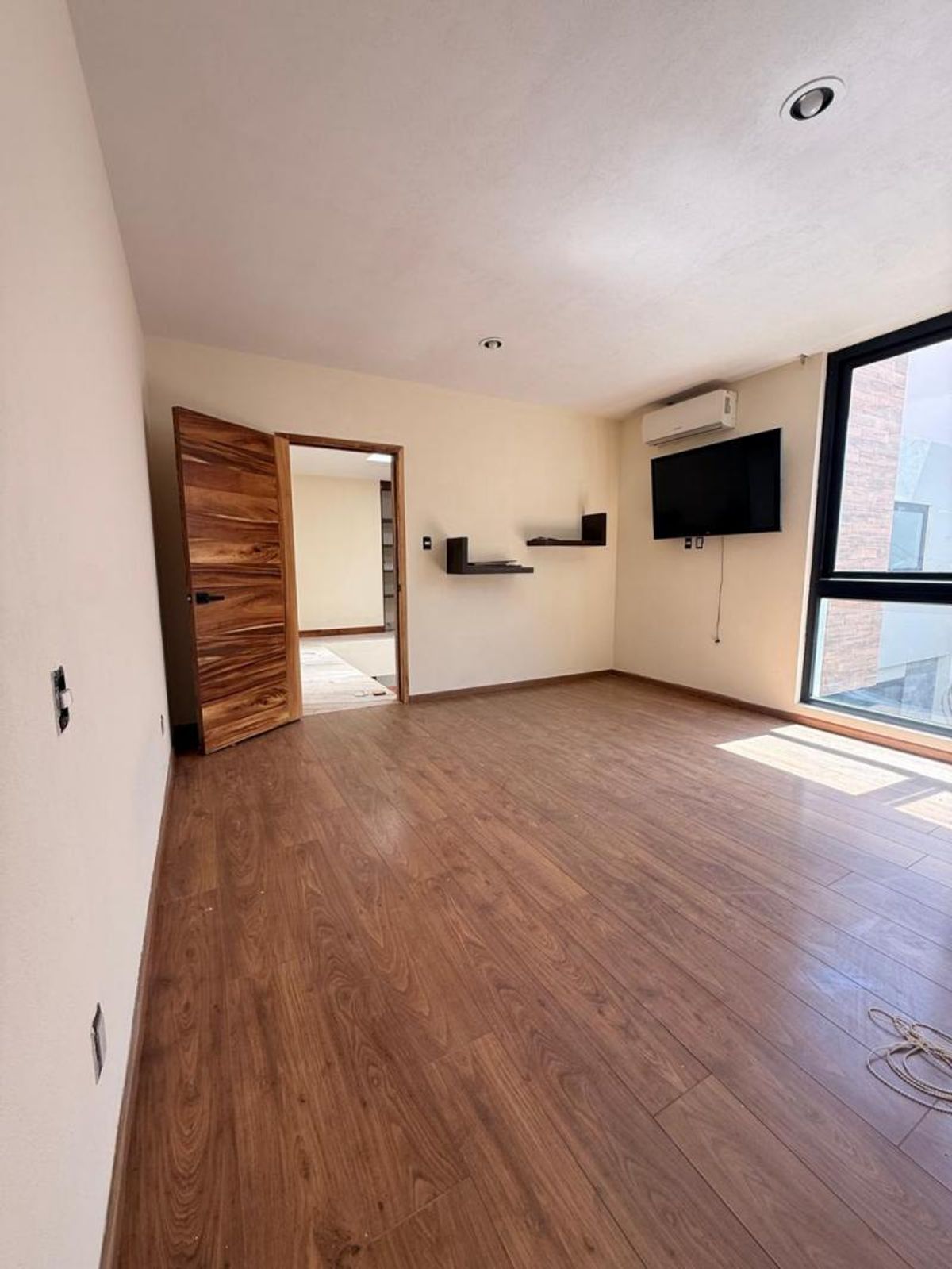 Habitación vacía con piso laminado de madera, puerta abierta, repisas flotantes y gran ventanal
