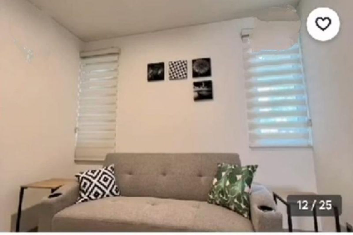 Sofá gris con cojines entre dos ventanas con persianas blancas y cuadros decorativos